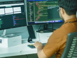 एआई एजेन्ट बढ्दै जाँदा सुरक्षा र निगरानी सीप किन महत्त्वपूर्ण? A person working at two computer monitors, representing AI agent operations and IT systems.